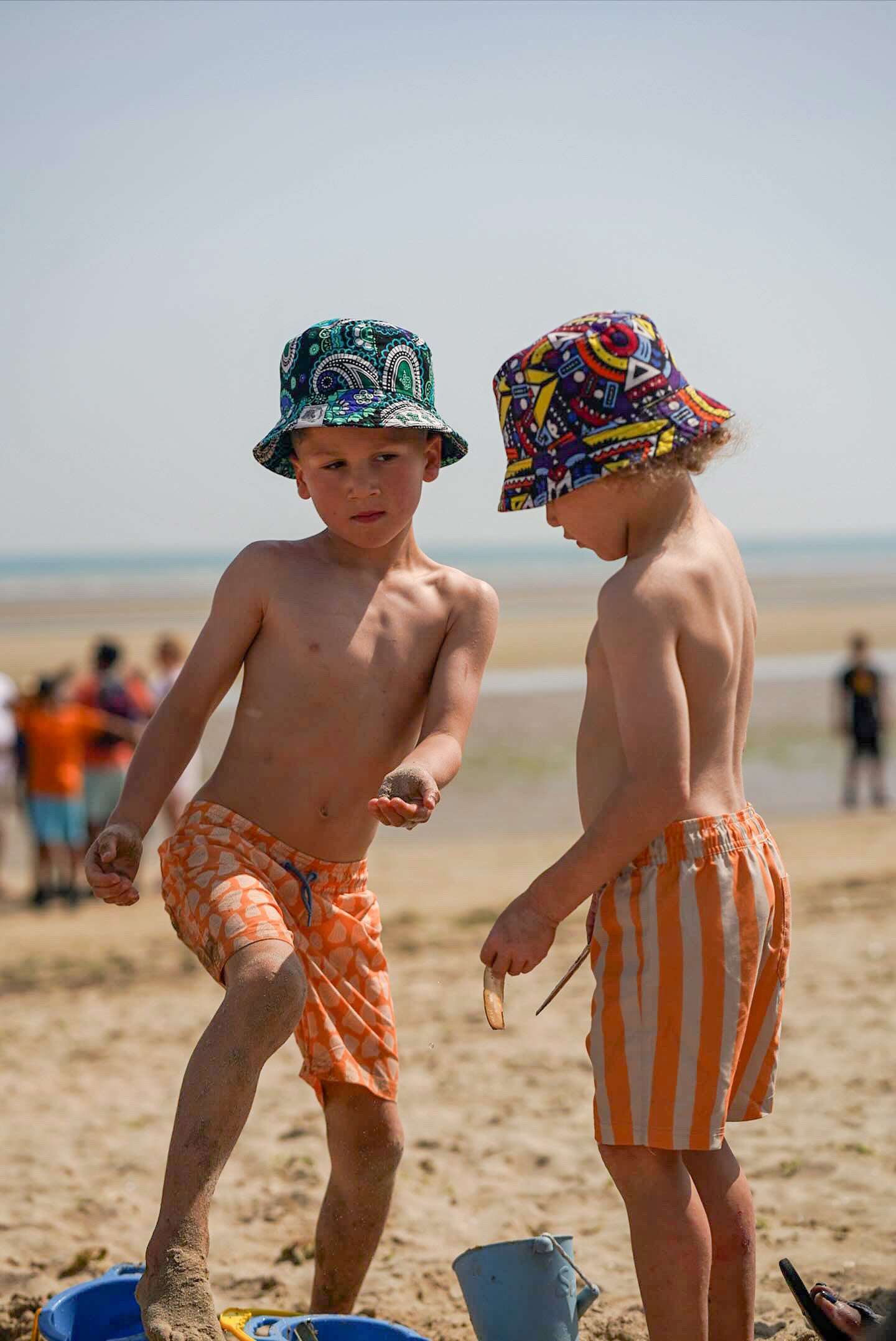 Two children on a beach wearing colorful Ben's Originals hats and swim trunks.