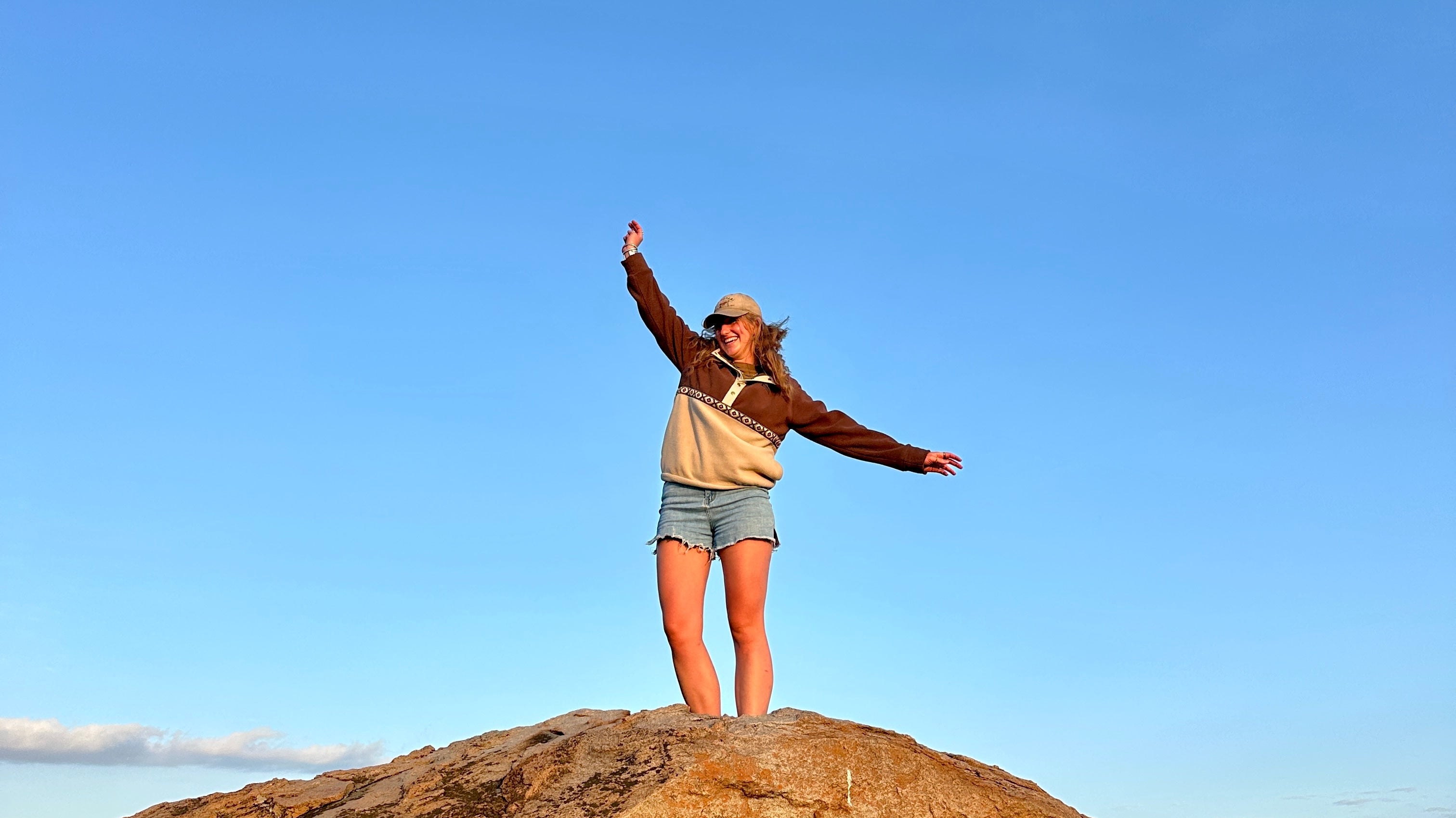 Person standing on a rocky outcrop with arms outstretched against a clear blue sky.